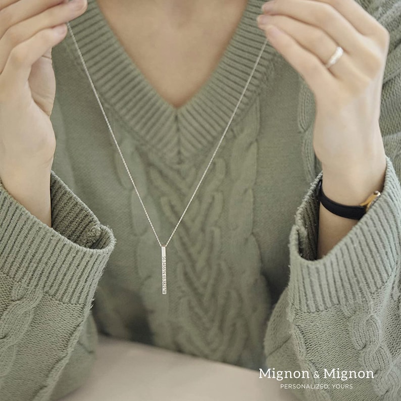 May include: A silver necklace with a long, thin pendant set with small, clear stones. The pendant is a vertical bar shape. The necklace is worn by a person wearing a green sweater.