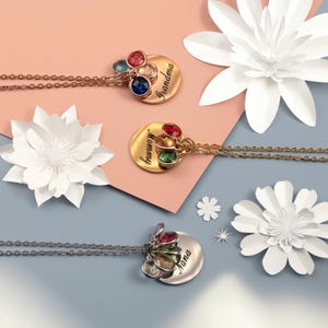 May include: Three gold and silver necklaces with charms. Each necklace has a round pendant with a different name engraved on it: "Grandma", "Mommy", and "Nana". Each pendant has five colorful gemstones attached.
