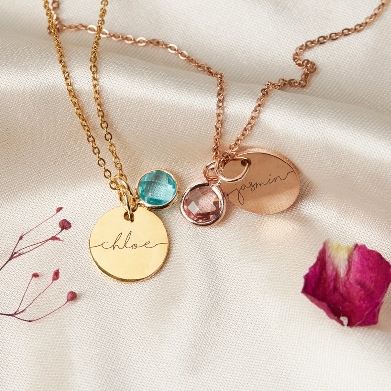 May include: Two gold necklaces with personalized name tags and small gemstone charms. The necklace on the left has a blue gemstone and a tag that says "chloe". The necklace on the right has a pink gemstone and a tag that says "jasmin".