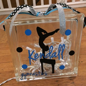 May include: A clear glass block with a silhouette of a gymnast on a balance beam, the name "Kendall" and the word "gymnastics" written in blue, and blue and black polka dots. The block is lit with white fairy lights.