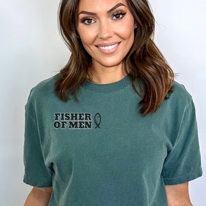 Puede incluir: Una camiseta verde oscuro con el texto "FISHER OF MEN" y un s&iacute;mbolo de pez.