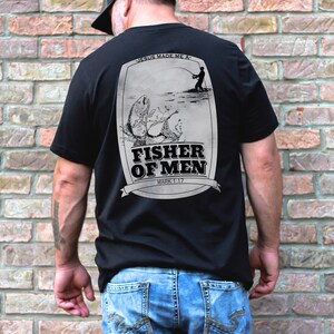 Puede incluir: Camiseta negra con un gr&aacute;fico de un pescador lanzando una l&iacute;nea y un pez saltando fuera del agua. El gr&aacute;fico est&aacute; rodeado por un borde blanco e incluye el texto "Jes&uacute;s me hizo un... Pescador de hombres Marcos 1:17".