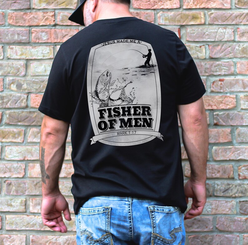 Puede incluir: Camiseta negra con un gr&aacute;fico de un pescador lanzando una l&iacute;nea y un pez saltando fuera del agua. El gr&aacute;fico est&aacute; rodeado por un borde blanco e incluye el texto "Jes&uacute;s me hizo un... Pescador de hombres Marcos 1:17".