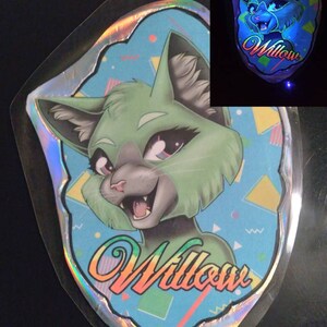 May include: A holographic sticker featuring a cartoon cat with green fur and gray accents. The cat has a friendly expression and the name "Willow" in a colorful, stylized font. The background is light blue with geometric shapes.