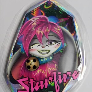 May include: A holographic sticker featuring a cartoon character with pink hair and a winking expression. The character has a bell collar and the word "Starfire" in pink block letters. The background shows a colorful space scene.