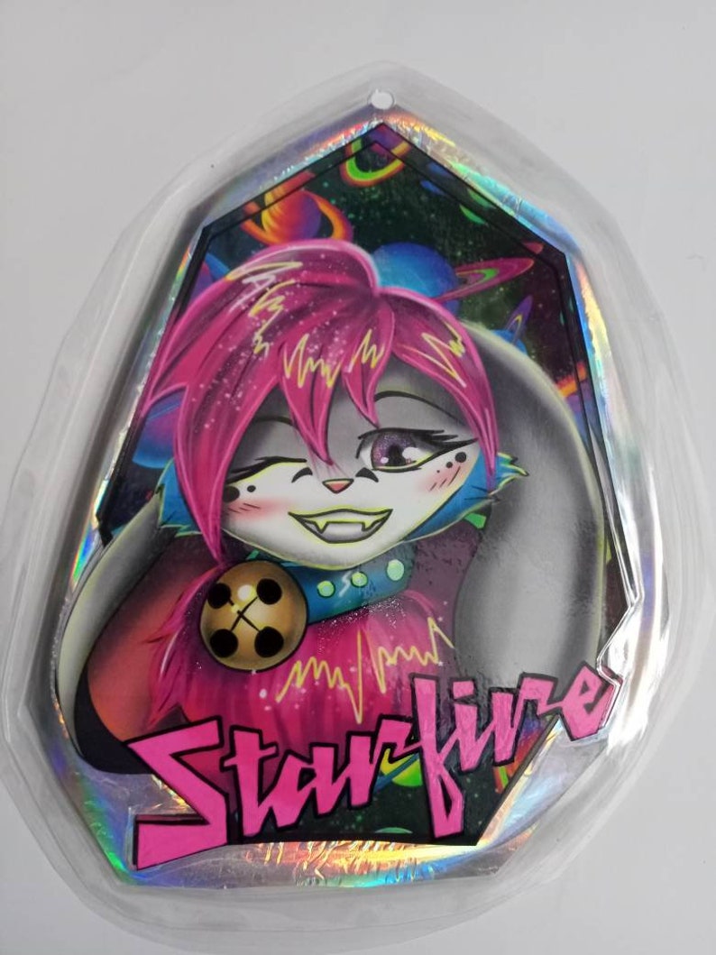 May include: A holographic sticker featuring a cartoon character with pink hair and a winking expression. The character has a bell collar and the word "Starfire" in pink block letters. The background shows a colorful space scene.