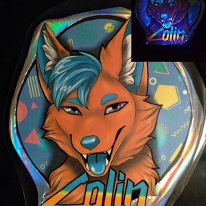 May include: A holographic sticker featuring a cartoon fox with orange fur, blue hair, and a blue tongue. The sticker has the name "Zolin" in colorful text. The background is light blue with geometric shapes.