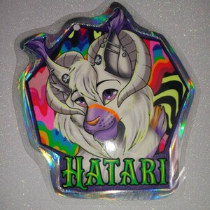 May include: A holographic charm featuring a stylized creature with white fur, purple accents, and large curved horns. The background has a psychedelic design with vibrant colors. The word "HATARI" is written in green letters.