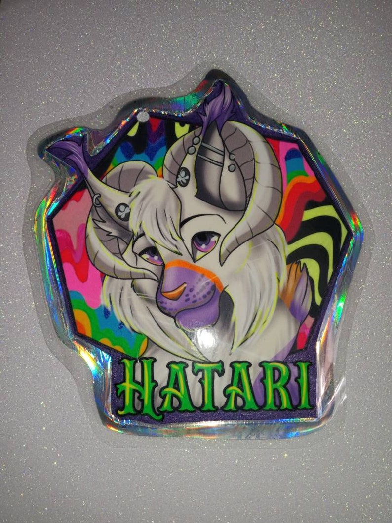 May include: A holographic charm featuring a stylized creature with white fur, purple accents, and large curved horns. The background has a psychedelic design with vibrant colors. The word "HATARI" is written in green letters.