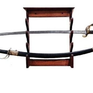 May include: A vintage sword and scabbard displayed on a wooden stand. The sword has a silver blade and a gold-colored hilt. The scabbard is black with gold accents. The stand is made of dark wood.