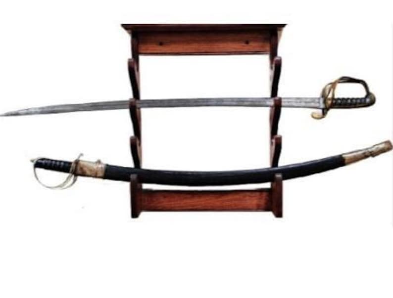 May include: A vintage sword and scabbard displayed on a wooden stand. The sword has a silver blade and a gold-colored hilt. The scabbard is black with gold accents. The stand is made of dark wood.
