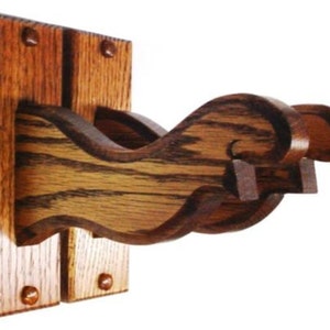 May include: A wooden wall-mounted gun rack with a dark brown finish. The rack features a decorative, curved design and is attached to a rectangular wooden base with four visible mounting pegs.