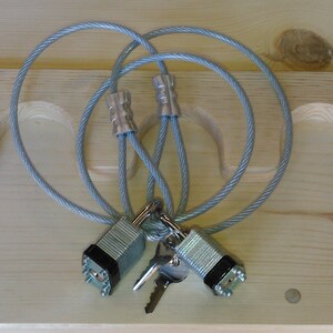 Puede incluir: Dos candados de combinaci&oacute;n plateados con llaves adjuntas a un cable de bloqueo gris. El cable est&aacute; en bucle y conectado con engarces plateados. Las cerraduras est&aacute;n sobre una superficie de madera clara.