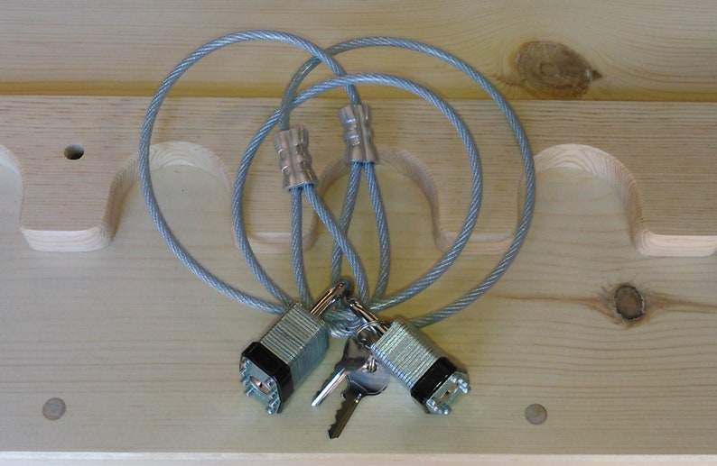 Puede incluir: Dos candados de combinaci&oacute;n plateados con llaves adjuntas a un cable de bloqueo gris. El cable est&aacute; en bucle y conectado con engarces plateados. Las cerraduras est&aacute;n sobre una superficie de madera clara.