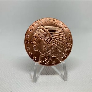 May include: A copper-colored coin featuring a profile of a Native American person wearing a headdress, with the word "LIBERTY" at the top. The coin is displayed on a clear acrylic stand.