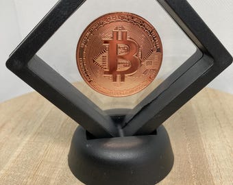 May include: A copper-colored Bitcoin coin displayed in a black, floating frame. The coin is centered within the frame, showcasing the Bitcoin logo and surrounding text. The frame is supported by a black base, set against a light wood-grain surface.