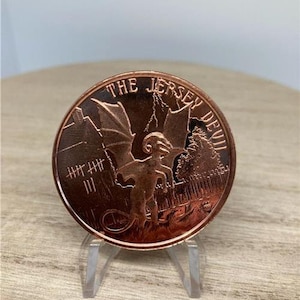 May include: A copper-colored round coin with a raised image of a winged creature with a tail and horns. The coin has the text "THE JERSEY DEVIL" around the edge.