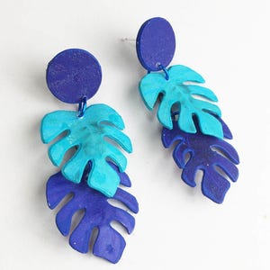May include: A pair of dangle earrings featuring a circular stud top in deep blue, connected to two layered monstera leaf shapes. The top leaf is a bright turquoise, while the bottom leaf is a deep blue, both crafted with a textured finish.