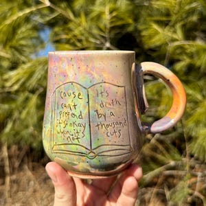 May include: A ceramic mug with a colorful, iridescent glaze. The mug features a book design with the text "Cause I can't pretend it's okay when it's not" and "It's death by a thousand cuts". The handle has an orange and yellow gradient.