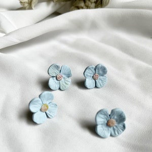 Peut inclure: Quatre boucles d'oreilles en forme de fleur bleu clair avec un centre rose clair. Les boucles d'oreilles sont sur une surface blanche.