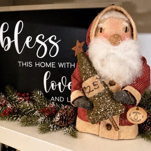 May include: A fabric Santa Claus figurine wearing a red and white checkered coat, holding a textured green tree with the number 25 and a round Merry Christmas tag. Behind it is a black sign with white text reading bless THIS HOME WITH love AND L