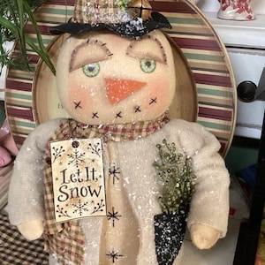 May include: A fabric snowman figurine wearing a black top hat with green sprigs and a jingle bell, a plaid scarf, and a beige coat. The snowman has an orange carrot nose, stitched facial features, and a wooden tag that reads Let It Snow.