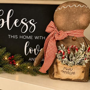 May include: A brown fabric gingerbread person figurine with a red and white gingham bow, featuring a small pocket with the text Merry Christmas, holding frosted greenery and candy canes, placed next to a sign reading bless THIS HOME WITH love.
