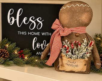 May include: A brown fabric gingerbread person figurine with a red and white gingham bow, featuring a small pocket with the text Merry Christmas, holding frosted greenery and candy canes, placed next to a sign reading bless THIS HOME WITH love.