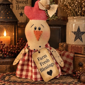 May include: A fabric chicken figurine with a red comb, orange beak, and black eyes. It wears a red and white checkered dress and a tan bow with a small bell. A rectangular tag hangs from the front, printed with the text Farm Fresh Blessings.