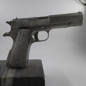 May include: A metallic grey handgun with a textured grip and a trigger. The firearm is displayed on a dark, rectangular base against a white background. The gun's design includes a slide, hammer, and sights.