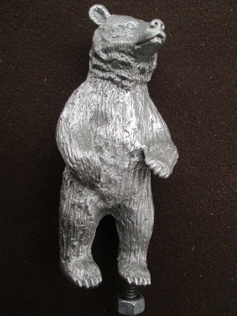 May include: A silver metal bear figurine standing on a metal screw. The bear has a textured surface and is standing on its hind legs.