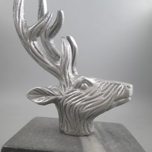 Puede incluir: Escultura de cabeza de ciervo de color plateado con astas detalladas. La escultura está sobre una base de mármol blanco y negro.