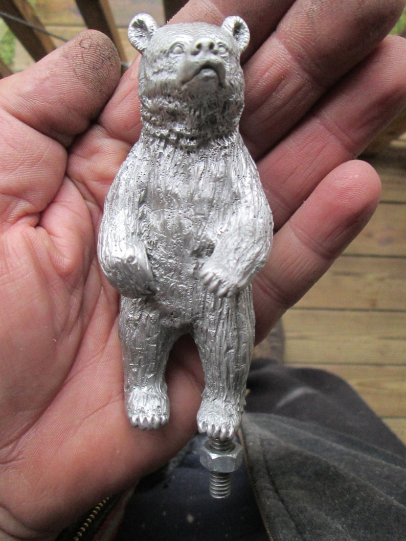 May include: A silver metal bear figurine standing on a metal base. The bear is standing on its hind legs with its paws raised. The bear has a detailed, textured surface.