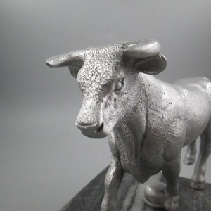 May include: A silver metal sculpture of a bull standing on a black marble base. The bull is facing forward with its head slightly tilted down. The sculpture is detailed with textured fur and a realistic depiction of the bull's features.