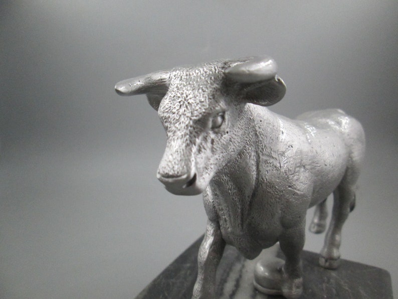 May include: A silver metal sculpture of a bull standing on a black marble base. The bull is facing forward with its head slightly tilted down. The sculpture is detailed with textured fur and a realistic depiction of the bull's features.