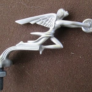 Könnte beinhalten: Silberfarbene geflügelte Figur mit einem Rad, einem stilisierten Design und einem Gewindesockel. Die Figur befindet sich in einer laufenden Pose mit ausgebreiteten Flügeln.