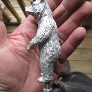 May include: A silver metal bear figurine with a textured surface. The bear is standing on a threaded metal rod with a nut.