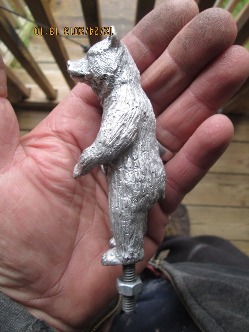May include: A silver metal bear figurine with a textured surface. The bear is standing on a threaded metal rod with a nut.