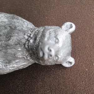 May include: A silver-colored bear figurine with a detailed, textured fur design. The bear has a realistic face with a small, black nose and dark eyes.