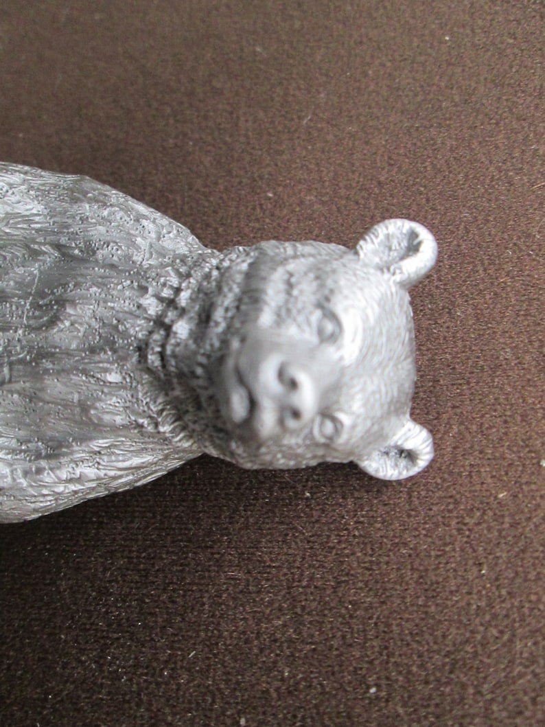 May include: A silver-colored bear figurine with a detailed, textured fur design. The bear has a realistic face with a small, black nose and dark eyes.