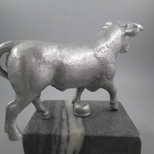 May include: A silver metal sculpture of a bull standing on a black and white marble base. The bull is facing to the right and has its head turned back to look over its shoulder.