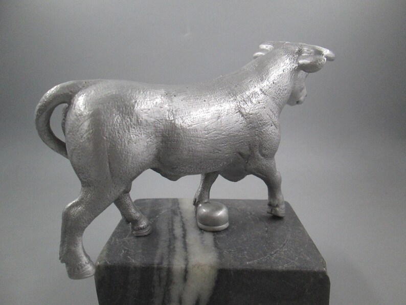 May include: A silver metal sculpture of a bull standing on a black and white marble base. The bull is facing to the right and has its head turned back to look over its shoulder.