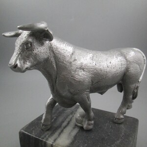May include: A silver metal bull figurine standing on a black and white marble base.