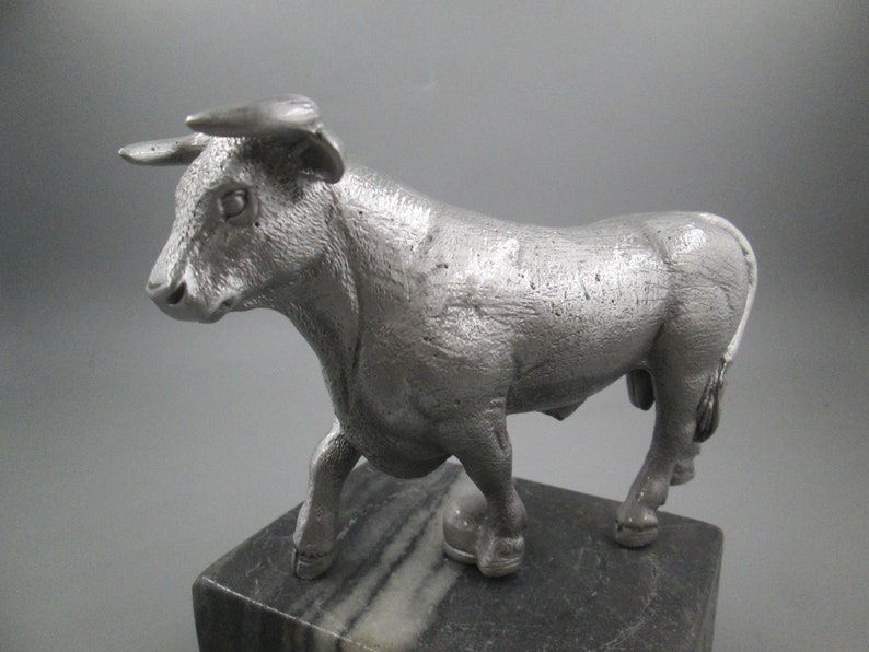 May include: A silver metal bull figurine standing on a black and white marble base.