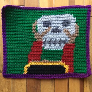 May include: A crocheted square with a skull design in shades of gray, brown, and red. The skull is surrounded by a green background with a purple border.
