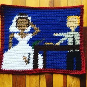 May include: Crocheted wall art depicting a bride and groom in a blue background with a table and chairs. The bride is wearing a white dress and the groom is wearing a white shirt and black pants.