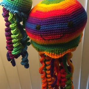 May include: Two crocheted rainbow jellyfish with smiling faces. The larger jellyfish has a multi-colored body and tentacles, while the smaller jellyfish has a green body and purple tentacles.