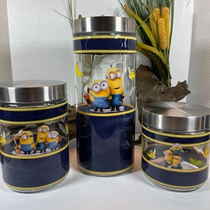 Op de afbeelding: Set van drie glazen potten met roestvrijstalen deksels. De potten hebben een blauw en geel ontwerp met een cartoonafbeelding van de Minions.