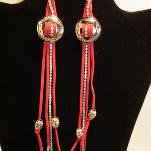 May include: A pair of long, red suede earrings with silver-tone accents. Each earring features a circular, silver-tone centerpiece with a red insert and rhinestone detailing. Dangling from the center are red suede strips, rhinestone chains, and silver-tone charms.