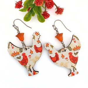 May include: A pair of dangle earrings featuring a wooden chicken charm with a white and orange chicken pattern. Each charm has a dangling orange flower bead.
